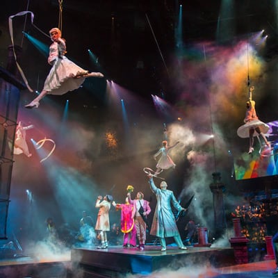 Coloridos personajes miran y señalan a mujeres vestidas de blanco que cuelgan de cuerdas en el aire - The Beatles Love Cirque du Soleil